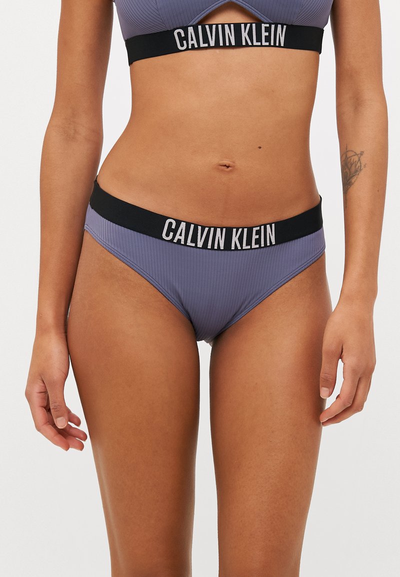 Vijolične bikini hlačke z črnim elastičnim pasom, ki ima belo blagovno znamko "CALVIN KLEIN". Zasnovane za tesno prileganje.