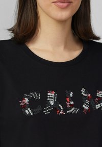 Zwarte t-shirt met een multikleurige pailletten tekstontwerp, met bloem- en geometrische patronen in rood, roze en wit. Rond de hals.