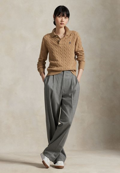 Nő áll zsebre tett kézzel, barna kötött pulóvert, szürke bő szárú nadrágot és fehér tornacipőt visel egyszínű háttér előtt.