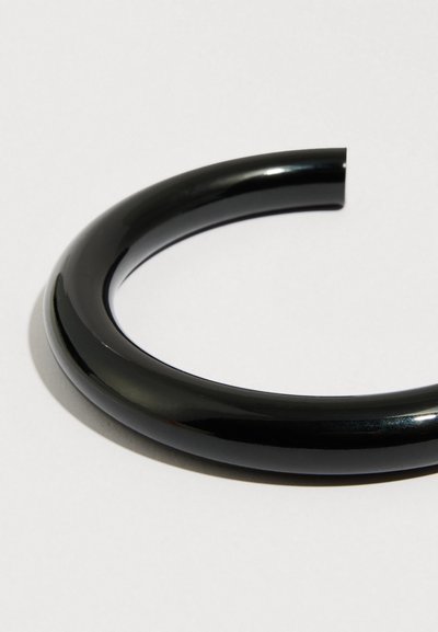 Musta kiiltävä rannekoru, jossa on pyöreä avoin muotoilu, sileällä pinnalla ja virtaviivaisilla, pyöristetyillä reunoilla. Ei näkyviä sulkimiä tai koristeita.