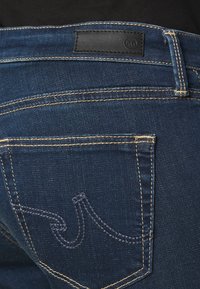 Tmavě modré džíny z denimu s kontrastním zlatým prošitím, obsahující koženou nášivku na pase a kapsový design se zakřiveným obrysem.
