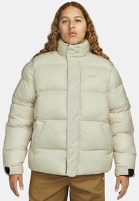 Beige péřová bunda s vysokým límcem, p�ředním zipem, dvěma bočními kapsami a malým černým logem na levé hrudi.