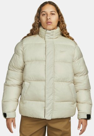 Beige puffy jakke med høj krave, med en frontlynlås, to sidelommer og et lille sort logo på venstre bryst.