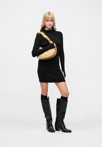 Rochie neagră cu tivuri, cizme negre până la genunchi și o geantă galbenă de umăr cu margini franjurate, prezentând un design modern, minimalist.