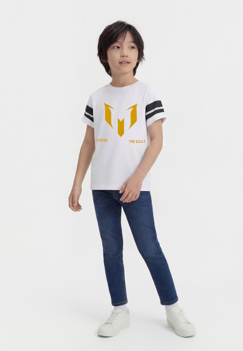 Αγόρι που στέκεται φορώντας λευκό μπλουζάκι με κίτρινο λογότυπο και κείμενο «LEGEND THE G.O.A.T.», μπλε τζιν και λευκά αθλητικά παπούτσια, κοιτώντας λίγο προς το πλάι.