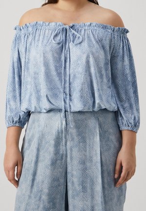 Kobieta ubrana w jasnoniebieską, wzorzystą bluzkę z odkrytymi ramionami i długimi rękawami oraz dopasowane luźne spodnie na jednolitym tle.