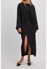 Mujer vistiendo un vestido negro de manga larga con mangas drapeadas transparentes y una gran apertura lateral, combinado con tacones negros de punta. 