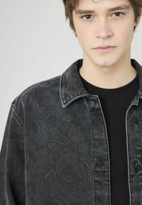 Veste en denim noir avec motifs texturés, fermeture à boutons et col classique, portée sur un t-shirt noir. Fond neutre.