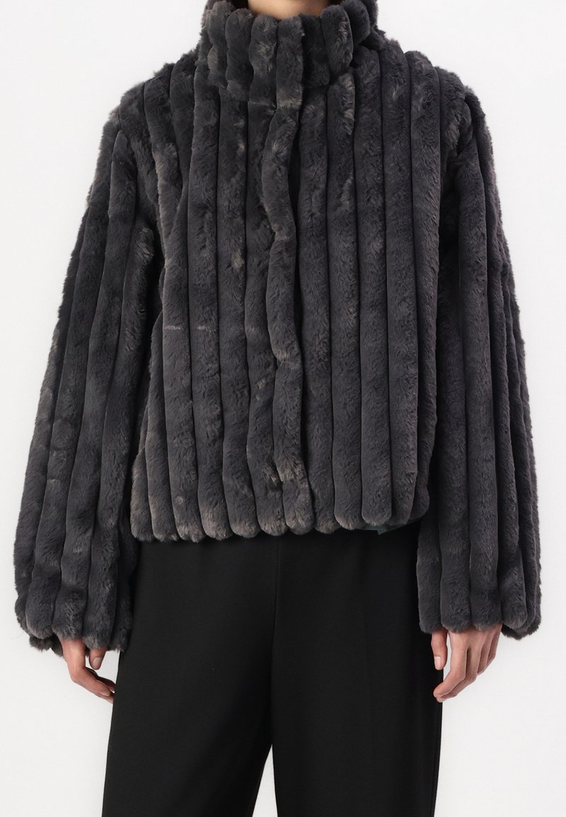 Personne portant une veste en fausse fourrure gris foncé à côtes verticales, manches longues et col montant, associée à un pantalon noir.