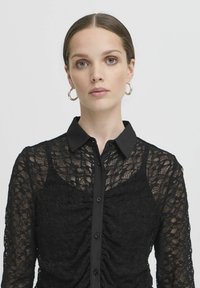 Blusa in pizzo nero con maniche lunghe, chiusura a bottoni frontale e colletto. Presenta una texture arricciata e un motivo floreale.