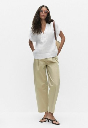 Kvinde med krøllet hår iført orange solbriller, hvid V-hals bluse, beige bukser med vide ben, sorte sandaler med hæl og med en skuldertaske i strå.