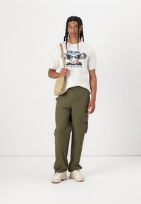 Casual outfit featuring a white graphic t-shirt with a bold design, olive green cargo pants, and beige sneakers. A tan crossbody bag is included.