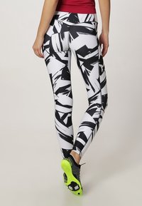 Persona que lleva leggings con un patrón en blanco y negro, una camiseta roja y zapatillas deportivas negras con suelas de un brillante amarillo-verde, de pie sobre una pierna.