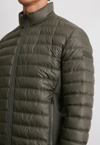 Oliwkowo-zielona pikowana kurtka z wysokim kołnierzem, poziomym pikowanym wzorem, zapięciem na zamek błyskawiczny z przodu oraz boczną kieszenią; gładka tekstura.