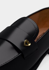 Γυαλιστερό μαύρο δερμάτινο μοκασίνι με ένα μικρό χρυσό μεταλλικό διακοσμητικό σε σχήμα καρδιάς στο λουράκι και ταμπά εσωτερική επένδυση.