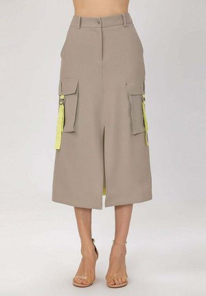 Jupe midi beige taille haute avec fente devant, deux grandes poches cargo latérales, détails de lanières jaunes, portée avec des talons à brides couleur nude.