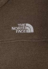 Brązowy polar z białym haftowanym logo "The North Face" na piersi, charakteryzujący się gładką teksturą i swobodnym stylem.
