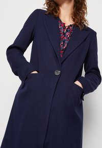 Marinblå kappa med skräddarsydd design, som har bred krage, enkel knappstängning och sidfickor. Slät textur och strukturerad form.