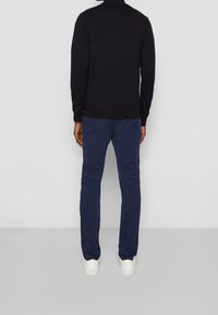 Hombre con una camiseta negra de manga larga, pantalones azul marino y zapatillas blancas de pie frente a una pared blanca sobre un suelo gris.