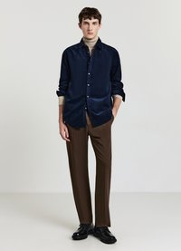 Camicia blu navy con bottoni e texture liscia, dolcevita beige sotto, pantaloni marroni e scarpe stringate nere. Design semplice e aderente.