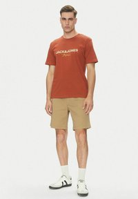 T-shirt di cotone color ruggine con logo bianco "JACK&JONES", abbinata a pantaloni beigi e sneakers bianche con accenti neri e calzini grigi.
