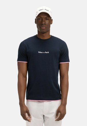 Homme portant un t-shirt bleu marine avec bordure rose et logo blanc "Eden Park", casquette blanche, et pantalon de couleur claire, debout devant un fond uni.
