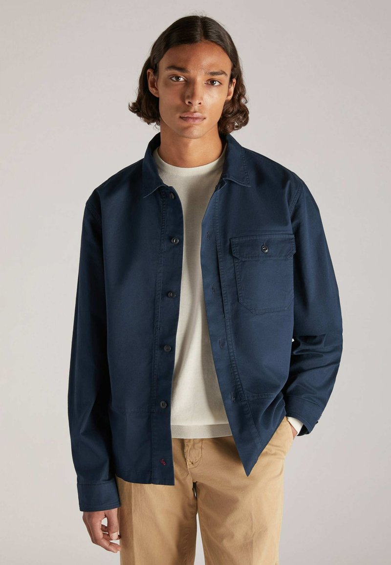 Un giovane con capelli ondulati lungo le spalle indossa una camicia blu navy a bottoni sopra un maglione chiaro e pantaloni khaki, in piedi contro uno sfondo neutro.