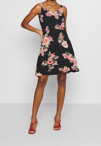 Robe noire à fleurs avec des roses roses, décolleté en V, et fines bretelles. Silhouette évasée tombant au-dessus du genou. Associée à des sandales à lanières rouges.