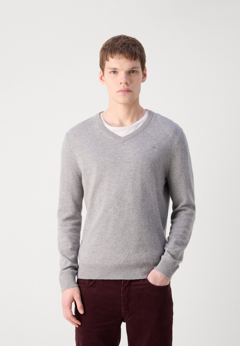 Helles graues V-Ausschnitt-Pullover aus weichem Material, mit langen Ärmeln und gerippten Bündchen. Getragen über einem weißen T-Shirt mit einem kleinen Logo.