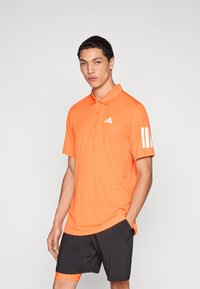 Polo naranja de tejido que absorbe la humedad, con cuello de dos botones, detalle de tres rayas blancas y corte ajustado. Combinado con pantalones cortos negros.