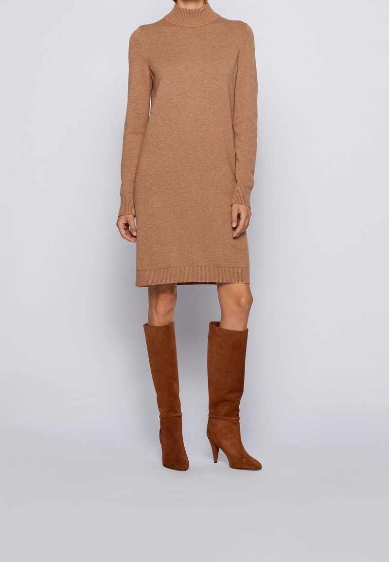 Femme portant une robe tricotée à manches longues de couleur camel et des bottes à talons en daim marron jusqu'aux genoux, se tenant devant un fond uni.