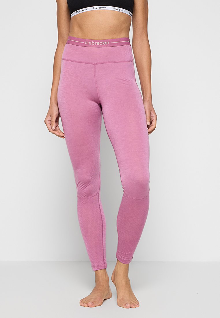 Nahaufnahme einer Person in hochgeschnittenen, rosa Icebreaker-Leggings und einem schwarzen, cropped Top, die barfuß vor einem schlichten Hintergrund steht.