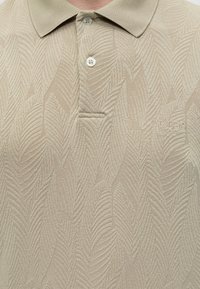 Μπεζ πόλο με υφή φύλλου, δύο λευκά κουμπιά στη λωρίδα και κέντημα "GUESS EST 1981" στο δεξί μέρος στο στήθος.