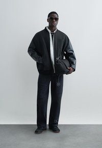 Jachetă bomber neagră cu mâneci din piele peste o cămașă albă, asortată cu blugi închiși la culoare cu accente cu nituri și pantofi negri. Accesoriile includ ochelari de soare.