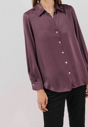 Frau trägt eine langärmlige lila Satinbluse mit Knöpfen und schwarze Hose, steht vor einem schlichten hellen Hintergrund.