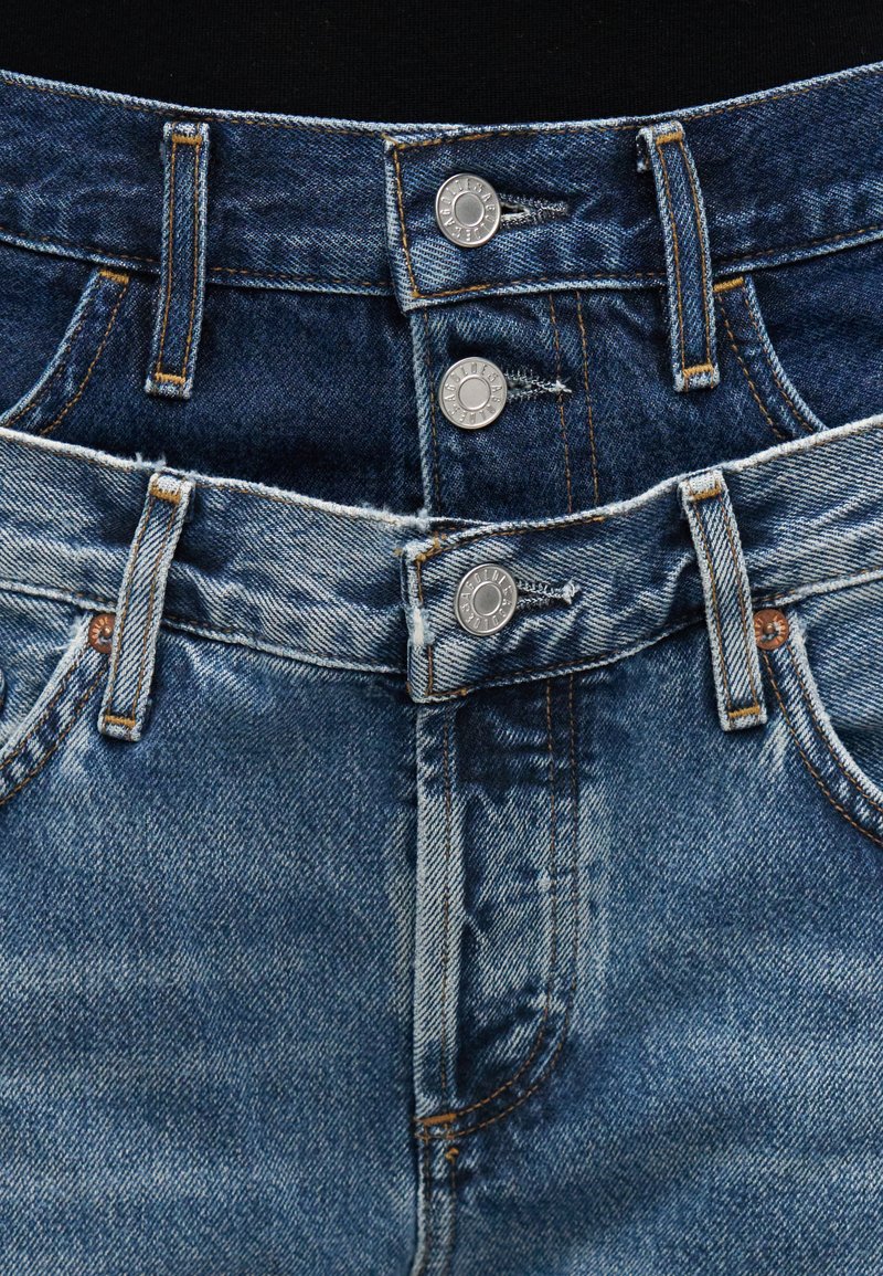 Jean en denim bleu avec une taille mi-haute, une fermeture à deux boutons, et des détails usés subtils le long des bords et des poches.
