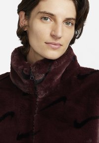 Burgundyröd fake fur-jacka med hög krage, dragkedjestängning och svarta abstrakta mönsterdetaljer, med en mjuk textur och avslappnad passform.