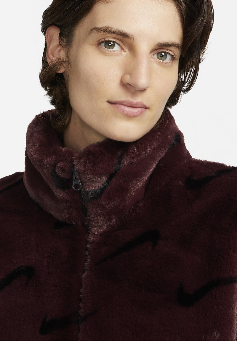 Gros plan sur une personne avec des cheveux bruns courts portant une veste en fausse fourrure bordeaux foncé à col montant avec des motifs noirs subtils.