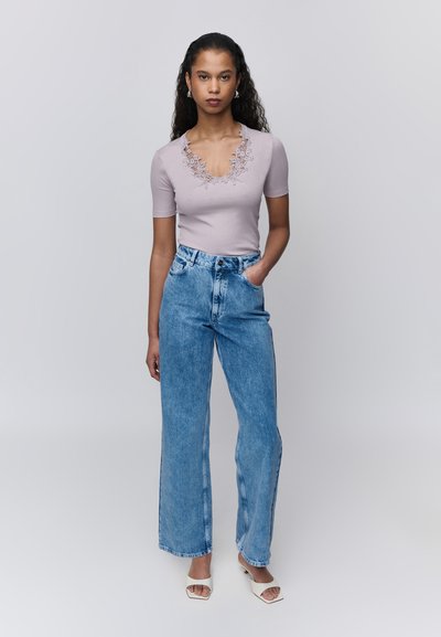 Femme debout, une main dans la poche, portant un haut à manches courtes violet clair bordé de dentelle, un jean bleu taille haute à jambes larges, et des talons blancs à bouts ouverts.