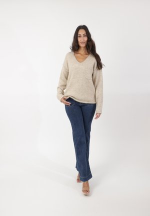 Pull en V en tricot beige avec une poche, associé à un jean flare bleu foncé et des sandales à talons transparents. Fond neutre.