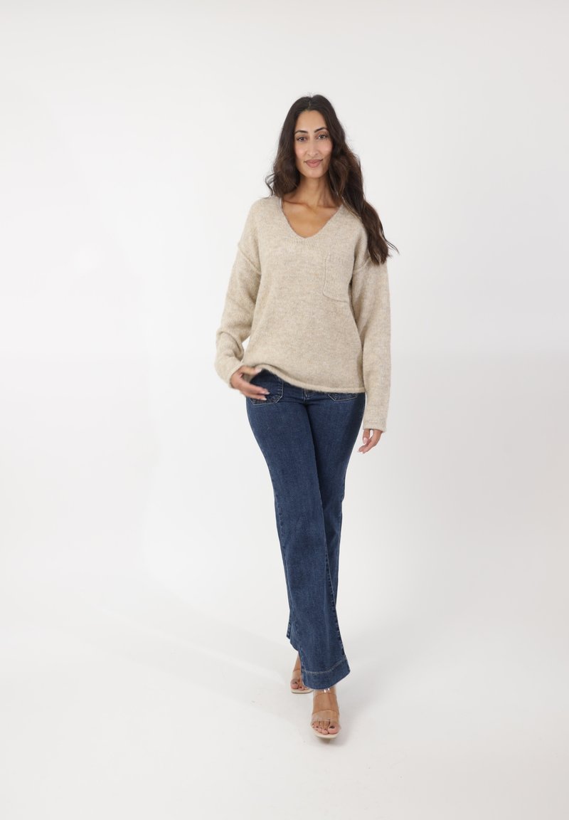 Beige, gestrickter V-Ausschnitt-Pullover mit einer Tasche, kombiniert mit dunkelblauen Schlagjeans und klaren High-Heel-Sandalen. Neutraler Hintergrund.