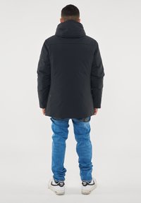 Giubbotto imbottito nero con colletto alto e cappuccio, caratterizzato da una texture liscia. Abbinato a jeans azzurri chiari e sneaker bianche con accenti neri.