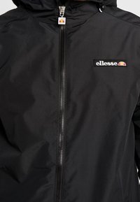 Sort vandtæt jakke med lynlås, der har hætte og et lille broderet logo på brystet. Glat tekstur og minimalistisk design.