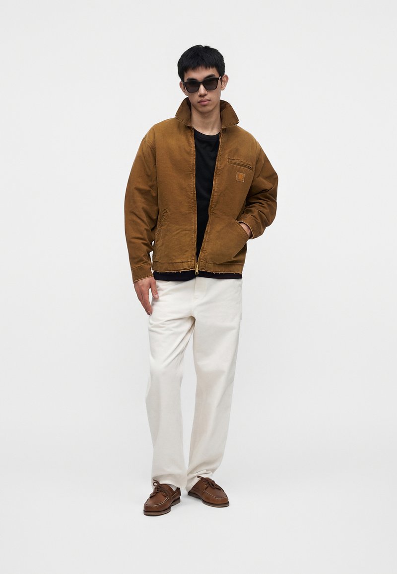 Mann mit schwarzen Sonnenbrillen, brauner Jacke, schwarzem Hemd, weißen Hosen und braunen Schuhen steht vor einem einfarbigen weißen Hintergrund.
