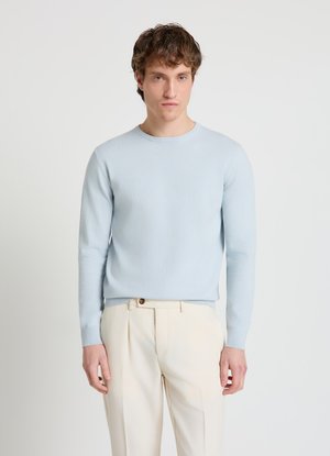Giovane uomo con capelli corti e ricci che indossa un maglione azzurro a girocollo e pantaloni beige plissettati su uno sfondo bianco semplice.