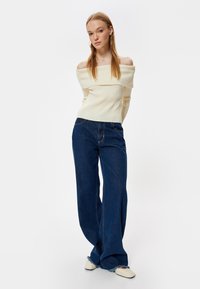 Rippiger, off-shoulder cremfarbener Pullover, kombiniert mit hoch taillierten, dunkelblauen weit geschnittenen Jeans mit subtilen Nähten und ausgefransten Säumen.