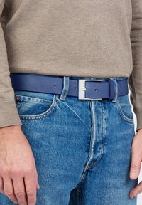 Tummansininen nahkavyö, jossa on hopeinen suorakulmainen solki, käytetty vaaleansinisien farkkujen päällä. Pintatekstuuri ja näkyvät ompeluyksityiskohdat.