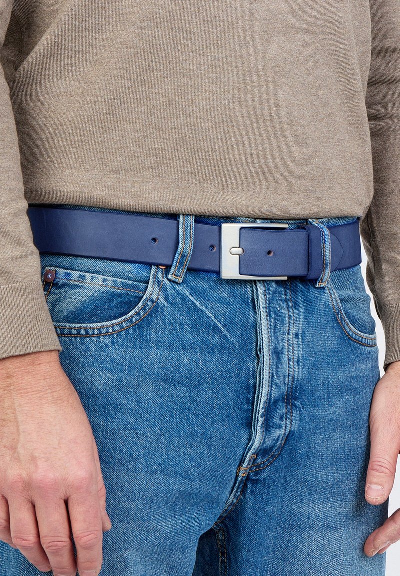 Tummansininen nahkavyö, jossa on hopeinen suorakulmainen solki, käytetty vaaleansinisien farkkujen päällä. Pintatekstuuri ja näkyvät ompeluyksityiskohdat.