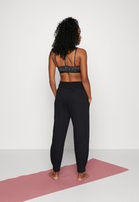 Žena s kudrnatými vlasy stojí bosá na růžové jóga podložce, na sobě má černou sportovní podprsenku s vzorem a volné černé kalhoty, proti bílému pozadí.