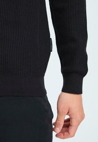 Schwarzer gerippter Pullover, der den strukturierten Stoff, enganliegende Bündchen und einen Bund zeigt. Ein Etikett mit "FRIDAY TRASH" ist sichtbar an der Seite.